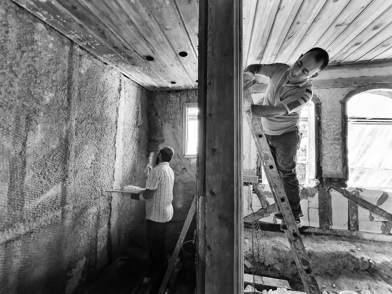services-03 Two men renovating a room, one on a ladder applying plaster, showcasing teamwork and effort.